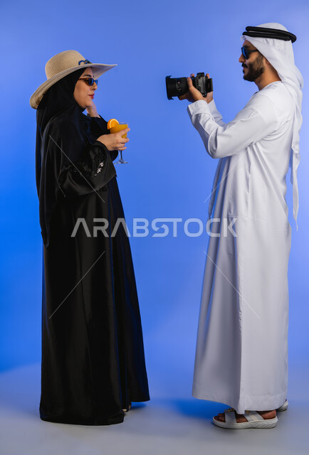 Drinking refreshing juices, having fun, family trip on summer vacation, portrait of an Arab Gulf Emirati couple wearing sunglasses, man taking pictures of his wife with the camera, full body, blue background.