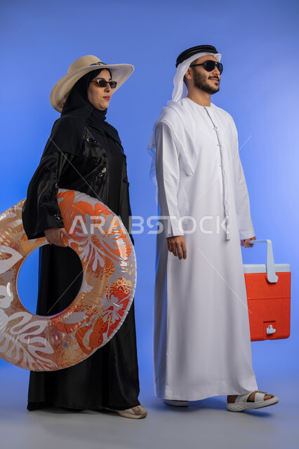 Preparing to go on a tour with expressions of joy and happiness, portrait of an Emirati Gulf Arab couple holding a float and a red water cooler in their hands, looking forward with happy gestures, having a good time in tourism and travel, full body, blue background.