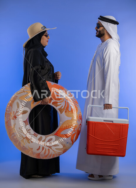 الاستعداد لقضاء إجازة صيفية ممتعة, بورتريه من الجانب لزوجين عربيين خليجيين اماراتيين متقابلان ينظران وجها لوجه  بتعابير الفرح والسرور، تحضير عوامة السباحة وصندوق تبريد المشروبات، الجسم بالطول الكامل، خلفية زرقاء.
