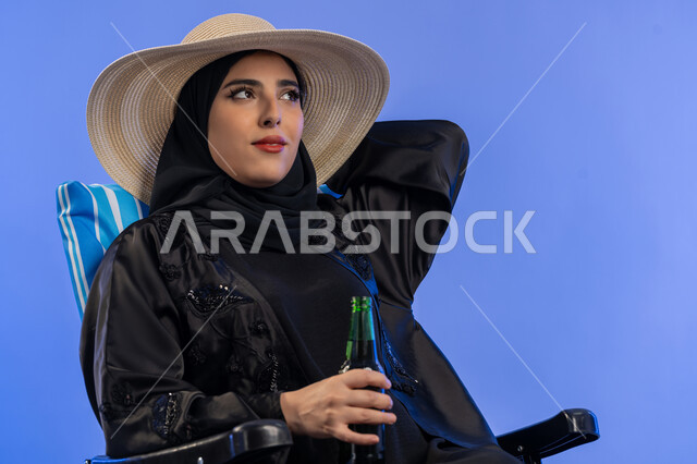 استرخاء واستجمام صيفي. بورتريه  مقرب لسيدة خليجية إماراتية محجبة ترتدي عباءة سوداء وقبعة شمسية تجلس على كرسي شاطئ, امراة سعوديه تمسك بزجاجة مشروب باردة تنظر الي شيء ما بتعابير هادئة ومسترخية، خلفية زرقاء.