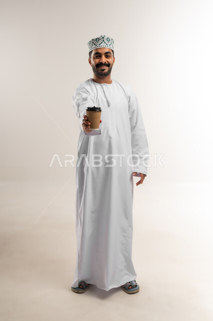 النظر الى الكاميرا بتعابير وايماءات السرور والسعاده, الاستمتاع بمذاق القهوه,  بورتريه لرجل عربي عماني خليجي يرتدي الدشداشه والكمه يحمل بيده كوب من المشروبات الساخنه, صورة الجسم بالطول الكامل, خلفيه بيضاء