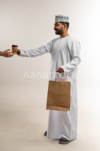 شحن الطلبات للزبائن، الاستفادة من عروض وتخفيضات الجمعة البيضاء، اقتناء المستلزمات والهدايا، بورتريه لرجل عربي خليجي عماني مبتسم يرتدي الدشداشه والكمه يحمل بيده أكياس تسوق ورقية  وكوب القهوة بإيماءات السرور، الجسم بالطول الكامل, خلفية بيضاء
