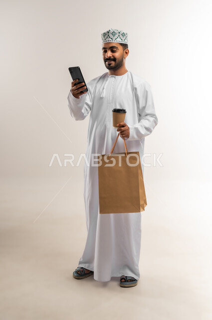 العمل على الهاتف المحمول، الاستفادة من عروض وتخفيضات الجمعة البيضاء، اقتناء المستلزمات والهدايا، بورتريه لرجل عربي خليجي عماني مبتسم يرتدي الدشداشه والكمه يحمل بيده أكياس تسوق ورقية  وكوب القهوة ينظر إلى شيء ما بإيماءات السرور، الجسم بالطول الكامل, خلفية بيضاء