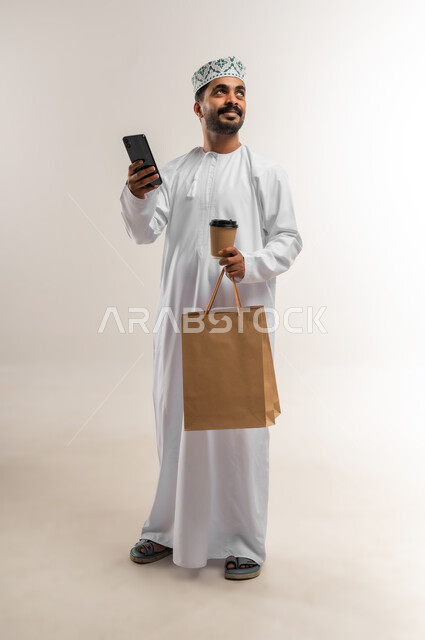 العمل على الهاتف المحمول، الاستفادة من عروض وتخفيضات الجمعة البيضاء، اقتناء المستلزمات والهدايا، بورتريه لرجل عربي خليجي عماني مبتسم يرتدي الدشداشه والكمه يحمل بيده أكياس تسوق ورقية  وكوب القهوة ينظر إلى شيء ما بإيماءات السرور، الجسم بالطول الكامل, خلفية بيضاء