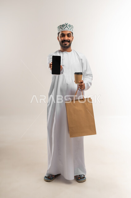 عرض تطبيقات الشراء والتبضع من الهاتف المحمول، الاستفادة من عروض وتخفيضات الجمعة البيضاء، اقتناء المستلزمات والهدايا، بورتريه لرجل عربي خليجي عماني مبتسم يرتدي الدشداشه والكمه يحمل بيده أكياس تسوق ورقية  وكوب القهوة ينظر إلى الكاميرا بإيماءات السرور والسعادة والحماس، الجسم بالطول الكامل, خلفية بيضاء