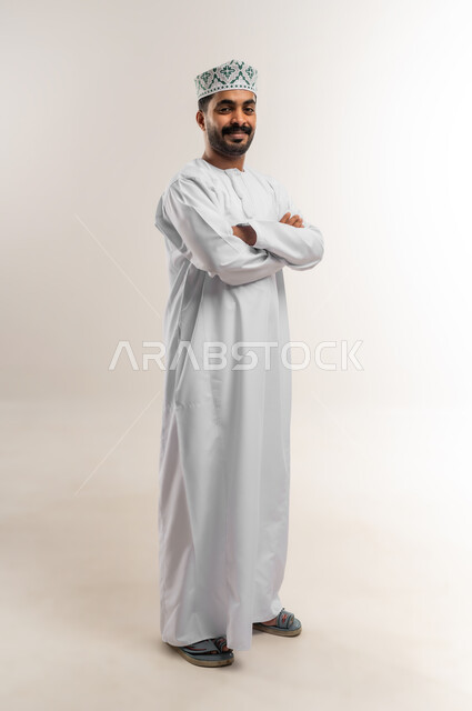 الاهتمام بالأناقة والمظهر الخارجي، مفهوم القوة والرجولة والشياكة، بورتريه لرجل عربي خليجي عماني يرتدي الدشداشه والكمة يقف بايدي متقاطعة ينظر الى الكاميرا بتعابير الفرح والسرور والثقة بالنفس، الجسم الكامل بالطول، خلفية بيضاء