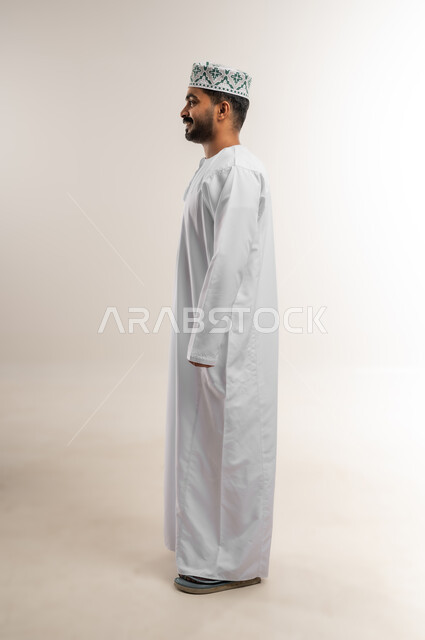 Concern for elegance and outward appearance, the concept of strength, masculinity and elegance, a side portrait of an Arab Gulf Omani man wearing a dishdasha and a kummah, looking somewhere with expressions of joy and pleasure, gestures of self-confidence, full body length, white background