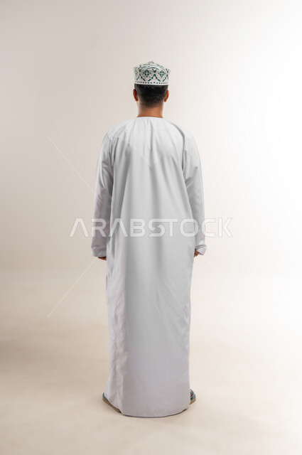 الاهتمام بالأناقة والمظهر الخارجي، مفهوم القوة والرجولة والشياكة، بورتريه من الخلف لرجل عربي خليجي عماني يرتدي الدشداشه والكمة يقف باستقامة بايماءات الثقة بالنفس، الجسم الكامل بالطول، خلفية بيضاء