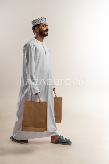 التسوق وطلب المنتجات عبر الإنترنت، توصيل الطلبات في الوقت المحدد وبشكل سليم، خدمة الشحن و التوصيل ذات الجودة العالية، بورتريه لشاب عربي خليجي عماني يرتدي الدشداشة والكمة يحمل بيده صندوق كرتوني مغلق، شراء الاحتياجات المنزلية، الجسم بالطول الكامل، خلفية بيضاء
