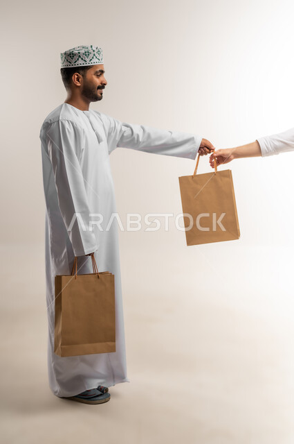 التسوق وطلب المنتجات عبر الإنترنت، توصيل الطلبات في الوقت المحدد وبشكل سليم، خدمة الشحن و التوصيل ذات الجودة العالية، بورتريه لشاب عربي خليجي عماني يرتدي الدشداشة والكمة يحمل بيده صندوق كرتوني مغلق، شراء الاحتياجات المنزلية، الجسم بالطول الكامل، خلفية بيضاء