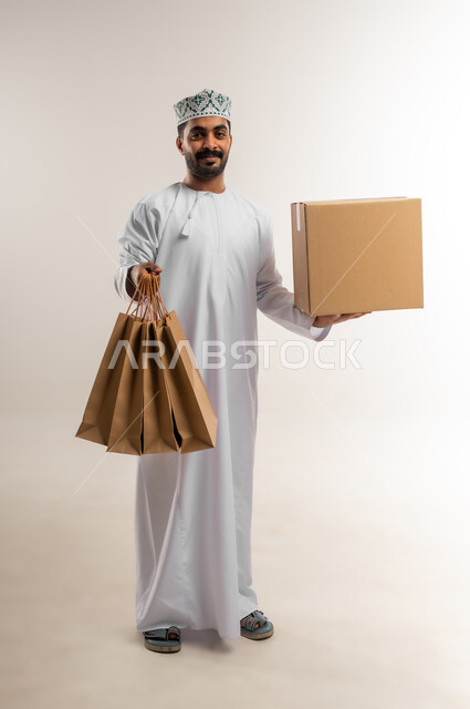 التسوق وطلب المنتجات عبر الإنترنت، توصيل الطلبات في الوقت المحدد وبشكل سليم، خدمة الشحن و التوصيل ذات الجودة العالية، بورتريه لشاب عربي خليجي عماني يرتدي الدشداشة والكمة يحمل بيده صندوق كرتوني مغلق، شراء الاحتياجات المنزلية، الجسم بالطول الكامل، خلفية بيضاء