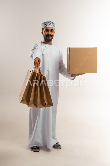 التسوق وطلب المنتجات عبر الإنترنت، توصيل الطلبات في الوقت المحدد وبشكل سليم، خدمة الشحن و التوصيل ذات الجودة العالية، بورتريه لشاب عربي خليجي عماني يرتدي الدشداشة والكمة يحمل بيده صندوق كرتوني مغلق، شراء الاحتياجات المنزلية، الجسم بالطول الكامل، خلفية بيضاء