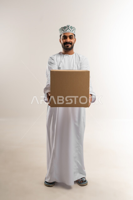التسوق وطلب المنتجات عبر الإنترنت، توصيل الطلبات في الوقت المحدد وبشكل سليم، خدمة الشحن و التوصيل ذات الجودة العالية، بورتريه لشاب عربي خليجي عماني يرتدي الدشداشة والكمة يحمل بيده صندوق كرتوني مغلق، شراء الاحتياجات المنزلية، الجسم بالطول الكامل، خلفية بيضاء