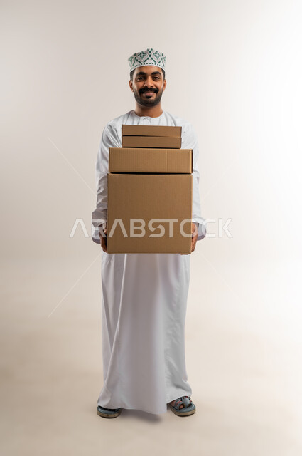 التسوق وطلب المنتجات عبر الإنترنت، توصيل الطلبات في الوقت المحدد وبشكل سليم، خدمة الشحن و التوصيل ذات الجودة العالية، بورتريه لشاب عربي خليجي عماني يرتدي الدشداشة والكمة يحمل بيده صندوق كرتوني مغلق، شراء الاحتياجات المنزلية، الجسم بالطول الكامل، خلفية بيضاء
