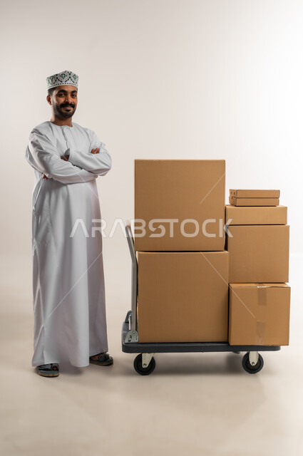On-time and accurate delivery of orders, high quality shipping and delivery service, portrait of a young Arab Gulf Omani man wearing a dishdasha and a kuma standing with his hands crossed beside closed cardboard boxes, shopping and ordering products online, full body, white background