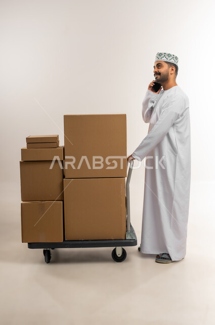Confirming reservations, managing express delivery services, answering calls and inquiries from customers, portrait of an Arab Gulf Omani man wearing a kummah and a dishdasha working in customer service at a freight and delivery company, documenting orders from a tablet, full-length body, white background