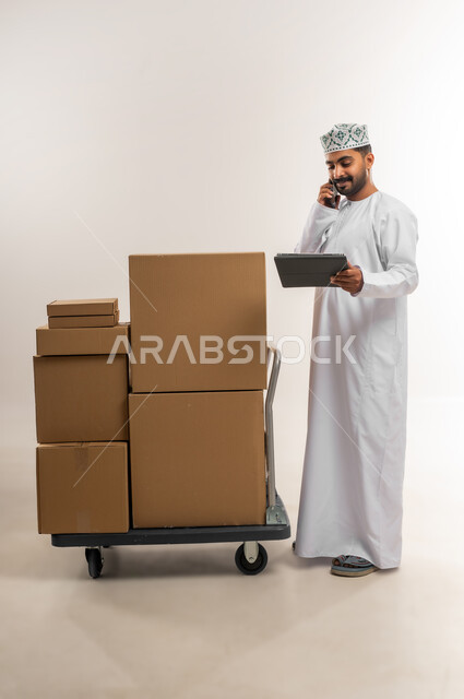 تاكيد الحجوزات ادارة عملية خدمات التوصيل السريع, الرد على الاتصالات والاستفسارات من الزبائن, بورتريه لرجل عربي خليجي عماني يرتدي الكمة والدشداشة يعمل في خدمة العملاء في شركة شحن وتوصيل البضائع, توثيق الطلبات من التابلت, الجسم بالطول الكامل، خلفية بيضاء