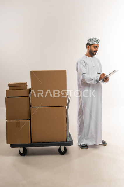 كتابة وتسجيل بيانات الشحنة، توصيل الطلبات في الوقت المحدد، خدمة الشحن و التوصيل ذات الجودة العالية، بورتريه لشاب عربي خليجي عماني يرتدي الدشداشة والكمة يقف بجانب بيده صناديق كرتونية مغلقة، التسوق وطلب المنتجات عبر الإنترنت، الجسم بالطول الكامل، خلفية بيضاء
