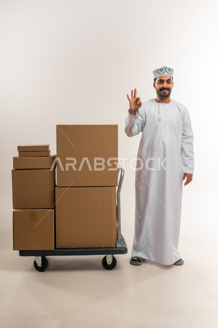 On-time delivery, high quality shipping and delivery service, portrait of a young Omani Gulf Arab man wearing a dishdasha and a kummah standing next to closed cardboard boxes, raising his hand with a quality and distinction sign, shopping and ordering products online, full body, white background