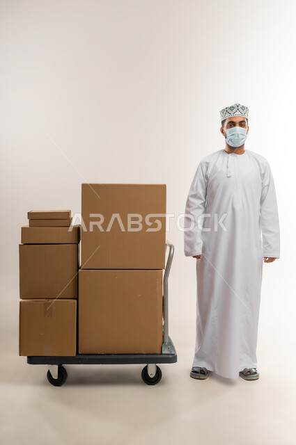 توصيل الطلبات في الوقت المحدد، خدمة الشحن و التوصيل ذات الجودة العالية، بورتريه لشاب عربي خليجي عماني يرتدي الدشداشة والكمة يضع الكمامة يقف بجانب بيده صناديق كرتونية مغلقة باستقامة، التسوق وطلب المنتجات عبر الإنترنت، الجسم بالطول الكامل، خلفية بيضاء