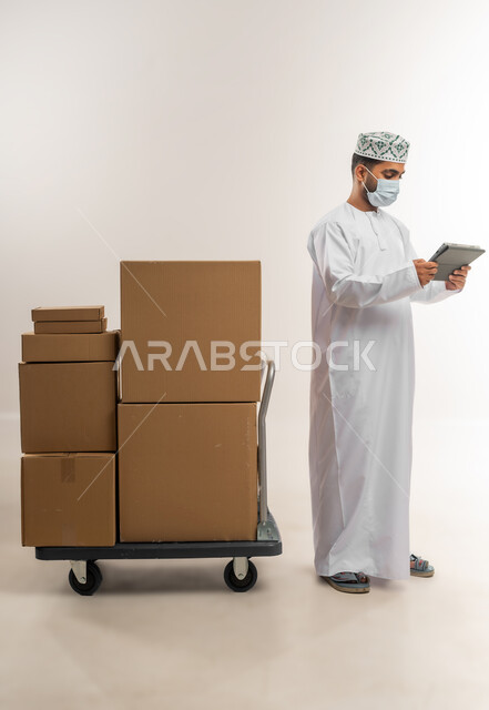 التسوق وطلب المنتجات عبر الإنترنت، توصيل الطلبات في الوقت المحدد وبشكل سليم، خدمة الشحن و التوصيل بجودة عالية، بورتريه لشاب عربي خليجي عماني يرتدي الدشداشة والكمة والكمامة يقف بجانب صناديق كرتونية، تسجيل الطلبات من جهاز التابلت، الجسم بالطول الكامل، خلفية بيضاء
