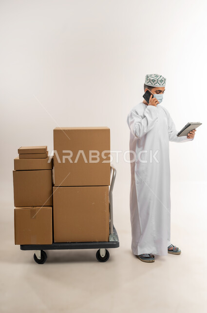 الرد على الاتصالات والاستفسارات, الالتزام بالاجراءات الاحترازية للوقاية من الامراض، بورتريه لرجل عربي خليجي عماني يرتدي الكمة والدشداشة يضع الكمامة يعمل في خدمة العملاء في شركة شحن وتوصيل البضائع, تاكيد الحجوزات وخدمات التوصيل السريع, الجسم بالطول الكامل، خلفية بيضاء
