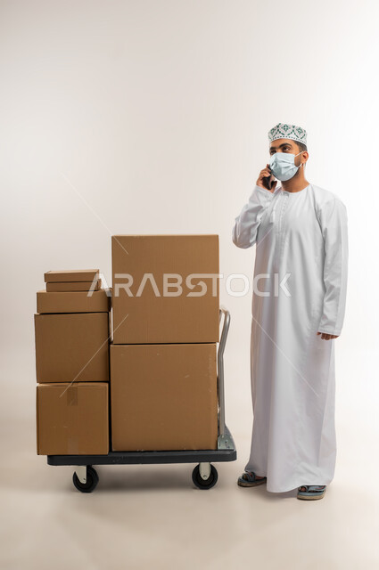 الرد على الاتصالات والاستفسارات, الالتزام بالاجراءات الاحترازية للوقاية من الامراض، بورتريه لرجل عربي خليجي عماني يرتدي الكمة والدشداشة يضع الكمامة يعمل في خدمة العملاء في شركة شحن وتوصيل البضائع, تاكيد الحجوزات وخدمات التوصيل السريع, الجسم بالطول الكامل، خلفية بيضاء