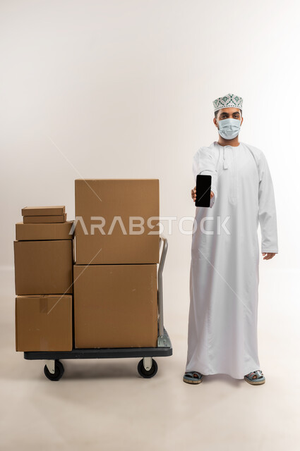 الالتزام بالاجراء الوقائي من الامراض، عرض تطبيق شراء المنتجات أونلاين, عمليات الدفع عن بعد, بورتريه لشاب عربي خليجي عماني يرتدي الدشداشة و الكمة والكمامة يقف بجانب صنايق كرتونية يعرض شاشاة سوداء فراغة عبر الهاتف المحمول، الجسم بالطول الكامل، خلفية بيضاء