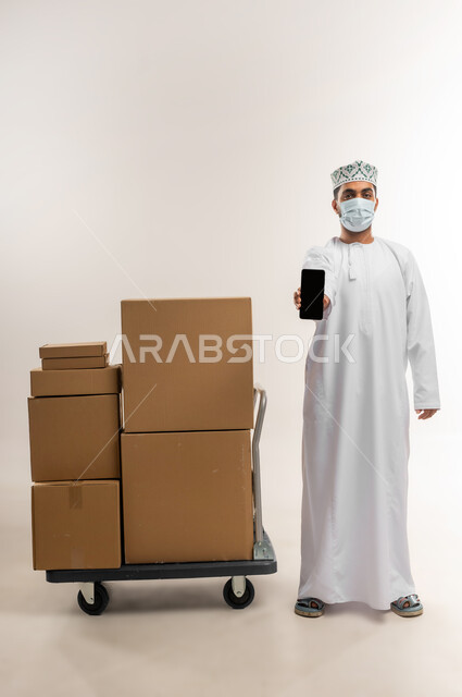 الالتزام بالاجراء الوقائي من الامراض، عرض تطبيق شراء المنتجات أونلاين, عمليات الدفع عن بعد, بورتريه لشاب عربي خليجي عماني يرتدي الدشداشة و الكمة والكمامة يقف بجانب صنايق كرتونية يعرض شاشاة سوداء فراغة عبر الهاتف المحمول، الجسم بالطول الكامل، خلفية بيضاء