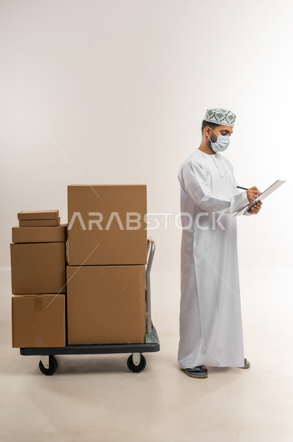 كتابة وتسجيل بيانات الشحنة، توصيل الطلبات في الوقت المحدد، خدمة الشحن و التوصيل ذات الجودة العالية، بورتريه لشاب عربي خليجي عماني يرتدي الدشداشة والكمة يقف بجانب بيده صناديق كرتونية مغلقة، التسوق وطلب المنتجات عبر الإنترنت، الجسم بالطول الكامل، خلفية بيضاء