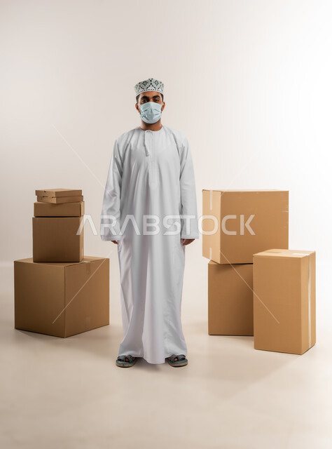 توصيل الطلبات في الوقت المحدد، خدمة الشحن و التوصيل ذات الجودة العالية، بورتريه لشاب عربي خليجي عماني يرتدي الدشداشة والكمة يضع الكمامة يقف بجانب بيده صناديق كرتونية مغلقة باستقامة، التسوق وطلب المنتجات عبر الإنترنت، الجسم بالطول الكامل، خلفية بيضاء