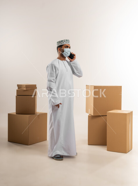 الالتزام بالاجراءات الاحترازية للوقاية من الامراض، الرد على الاتصالات والاستفسارات, بورتريه لرجل عربي خليجي عماني يرتدي الكمة والدشداشة يضع الكمامة يعمل في خدمة العملاء في شركة شحن وتوصيل البضائع, تاكيد الحجوزات وخدمات التوصيل السريع, الجسم بالطول الكامل، خلفية بيضاء