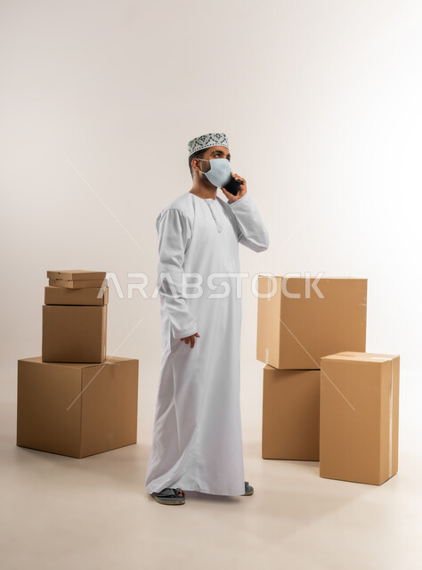 Adherence to precautionary measures to prevent diseases, responding to calls and inquiries, portrait of an Arab Gulf Omani man wearing a kuma and a dishdasha, wearing a mask, working in customer service at a freight and delivery company, confirming reservations and express delivery services, full-length body, white background