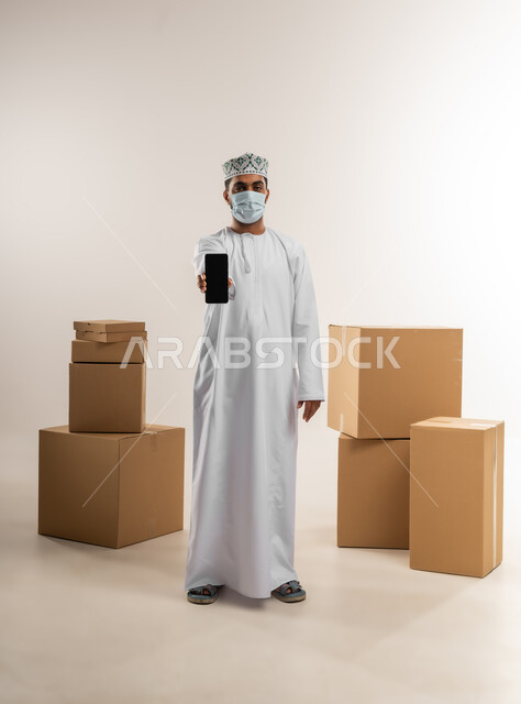الالتزام بالاجراء الوقائي من الامراض، عرض تطبيق شراء المنتجات أونلاين, عمليات الدفع عن بعد, بورتريه لشاب عربي خليجي عماني يرتدي الدشداشة و الكمة والكمامة يقف بجانب صنايق كرتونية يعرض شاشاة سوداء فراغة عبر الهاتف المحمول، الجسم بالطول الكامل، خلفية بيضاء