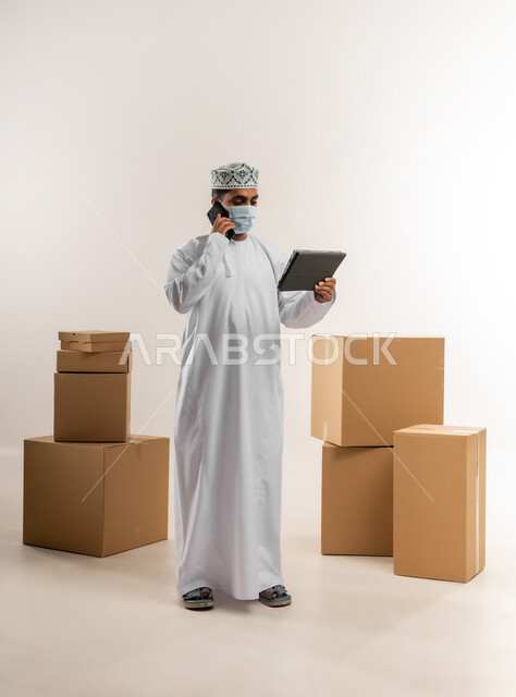الالتزام بالاجراءات الاحترازية للوقاية من الامراض، الرد على الاتصالات والاستفسارات, بورتريه لرجل عربي خليجي عماني يرتدي الكمة والدشداشة يضع الكمامة يعمل في خدمة العملاء في شركة شحن وتوصيل البضائع, تاكيد الحجوزات وخدمات التوصيل السريع, الجسم بالطول الكامل، خلفية بيضاء