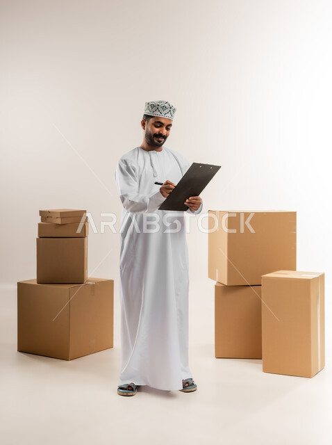 كتابة وتسجيل بيانات الشحنة، توصيل الطلبات في الوقت المحدد، خدمة الشحن و التوصيل ذات الجودة العالية، بورتريه لشاب عربي خليجي عماني يرتدي الدشداشة والكمة يقف بجانب بيده صناديق كرتونية مغلقة، التسوق وطلب المنتجات عبر الإنترنت، الجسم بالطول الكامل، خلفية بيضاء