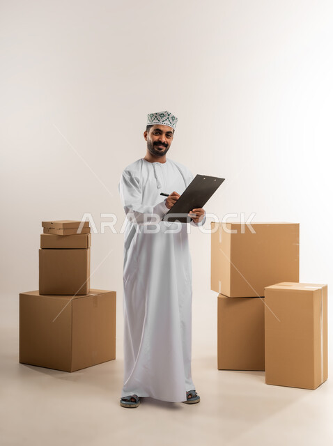 كتابة وتسجيل بيانات الشحنة، توصيل الطلبات في الوقت المحدد، خدمة الشحن و التوصيل ذات الجودة العالية، بورتريه لشاب عربي خليجي عماني يرتدي الدشداشة والكمة يقف بجانب بيده صناديق كرتونية مغلقة، التسوق وطلب المنتجات عبر الإنترنت، الجسم بالطول الكامل، خلفية بيضاء