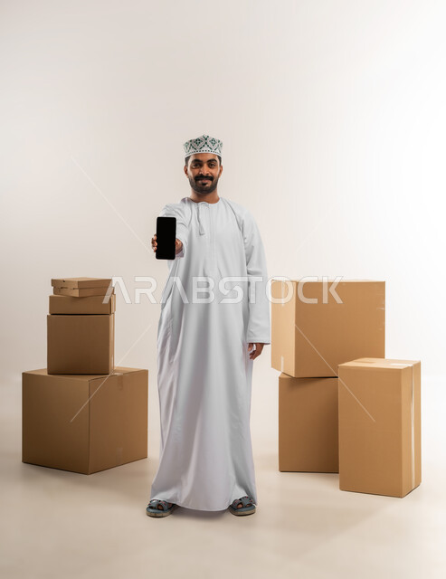 عرض تطبيقات شراء المنتجات أونلاين, عمليات الدفع عن بعد, مفهوم التسوق الالكتروني, بورتريه لشاب عربي خليجي عماني يرتدي الدشداشة و الكمة يفقف بجانب صنايق كرتونية يعرض شاشاة سوداء فراغة عبر الهاتف المحمول، الجسم بالطول الكامل، خلفية بيضاء