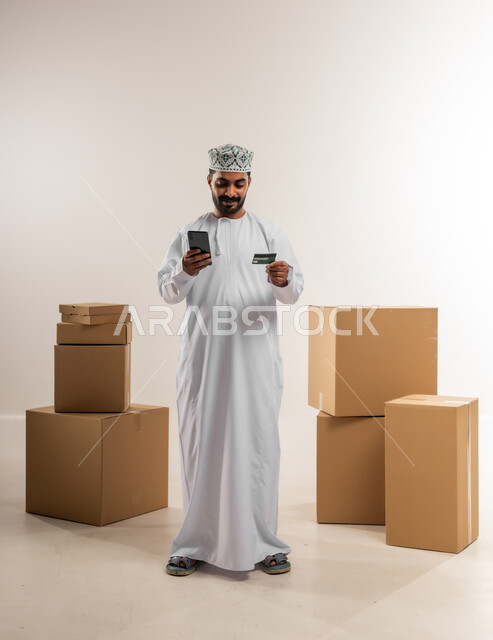 عمليات الدفع عن بعد, شراء المنتجات أونلاين, مفهوم التسوق الالكتروني, بورتريه لشاب عربي خليجي عماني يرتدي الدشداشة و الكمة يفقف بجانب صنايق كرتونية يمسك بيده الهاتف المحمول و بطاقة الائتمان البنكية, مفهوم خدمات التوصيل السريع, الجسم بالطول الكامل، خلفية بيضاء