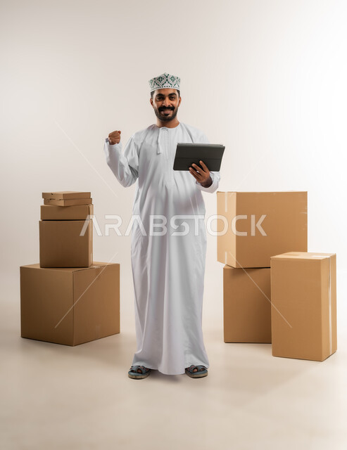 التسوق وطلب المنتجات عبر الإنترنت، توصيل الطلبات في الوقت المحدد وبشكل سليم، خدمة الشحن و التوصيل بجودة عالية، بورتريه لشاب عربي خليجي عماني يرتدي الدشداشة والكمة يقف بجانب صناديق كرتونية، تسجيل الطلبات من جهاز التابلت، الجسم بالطول الكامل، خلفية بيضاء