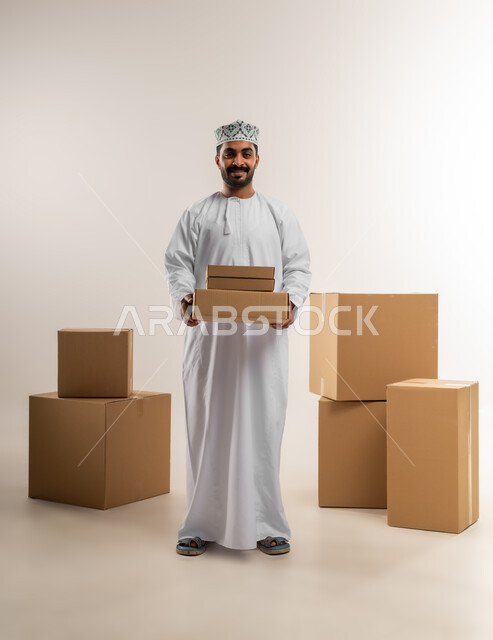 التسوق وطلب المنتجات عبر الإنترنت، توصيل الطلبات في الوقت المحدد وبشكل سليم، خدمة الشحن و التوصيل ذات الجودة العالية، بورتريه لشاب عربي خليجي عماني يرتدي الدشداشة والكمة يحمل بيده صندوق كرتوني مغلق، شراء الاحتياجات المنزلية، الجسم بالطول الكامل، خلفية بيضاء