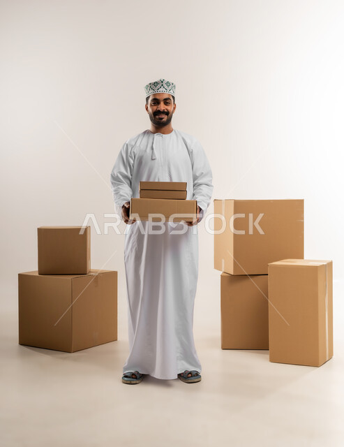التسوق وطلب المنتجات عبر الإنترنت، توصيل الطلبات في الوقت المحدد وبشكل سليم، خدمة الشحن و التوصيل ذات الجودة العالية، بورتريه لشاب عربي خليجي عماني يرتدي الدشداشة والكمة يحمل بيده صندوق كرتوني مغلق، شراء الاحتياجات المنزلية، الجسم بالطول الكامل، خلفية بيضاء