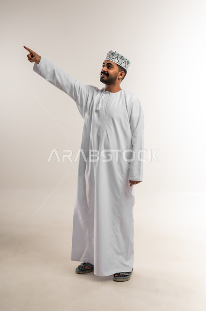 اشارات تحدد هذا المكان (هنا)، الاشارة إلى شيء ما، تعابير التحديد والتأكيد، بورتريه لرجل عربي خليجي عماني مبتسم يرتدي الدشداشه والكمة يشير بإصبع السبابة وينظر الى شيء ما بإيماءات السعادة والسرور، الجسم الكامل بالطول، خلفية بيضاء
