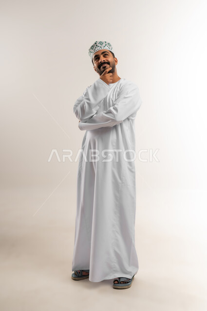 Deep thinking and absent-mindedness, trying to find a solution to difficult problems, anxiety and confusion in making the right decision, portrait of an Arab Gulf Omani man wearing a dishdasha and a kumrah, putting his hand under his chin and looking somewhere far away with gestures of integration, full-length body, white background