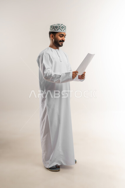 مراجعة نتائج الاجتماع، بورتريه لرجل عربي خليجي عماني يرتدي الدشداشة والكمة يحمل بيده كراسه تدوين الملاحظات يقوم بتصفح الاوراق، اعداد تقارير ومهام العمل، دراسة جدوى الاعمال، الجسم بالطول الكامل، خلفيه بيضاء