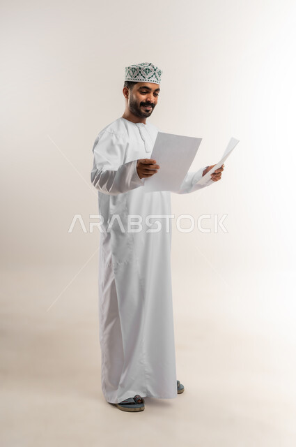 مراجعة نتائج الاجتماع، بورتريه لرجل عربي خليجي عماني يرتدي الدشداشة والكمة يحمل بيده كراسه تدوين الملاحظات يقوم بتصفح الاوراق، اعداد تقارير ومهام العمل، دراسة جدوى الاعمال، الجسم بالطول الكامل، خلفيه بيضاء