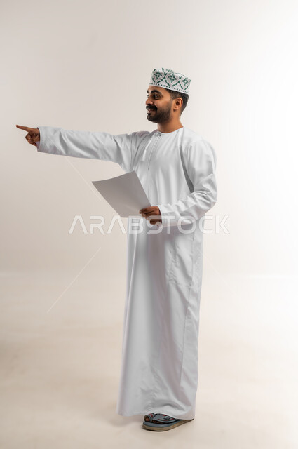 مراجعة نتائج الاجتماع، بورتريه لرجل عربي خليجي عماني يرتدي الدشداشة والكمة يحمل بيده كراسه تدوين الملاحظات يقوم بتصفح الاوراق، اعداد تقارير ومهام العمل، دراسة جدوى الاعمال، الجسم بالطول الكامل، خلفيه بيضاء