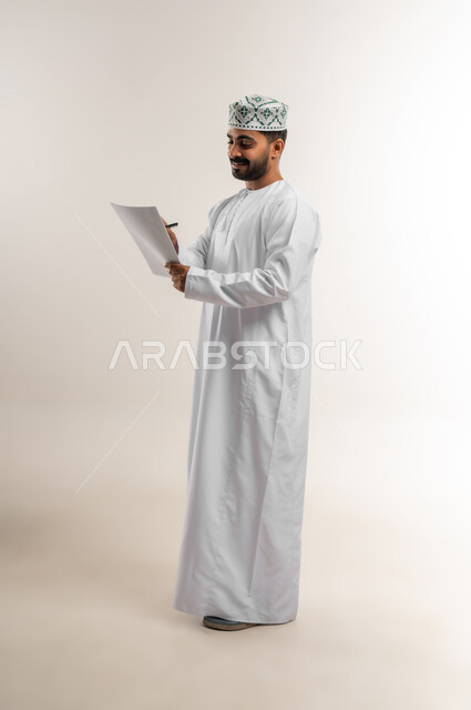 تدوين وكتابة التقارير، مراجعة نتائج الاجتماع، بورتريه لرجل عربي خليجي عماني يرتدي الدشداشة والكمة يحمل بيده كراسه تدوين الملاحظات يقوم بتصفح الاوراق، اعداد تقارير ومهام العمل، دراسة جدوى الاعمال، الجسم بالطول الكامل، خلفيه بيضاء