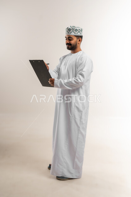 تدوين وكتابة التقارير، مراجعة نتائج الاجتماع، بورتريه لرجل عربي خليجي عماني يرتدي الدشداشة والكمة يحمل بيده كراسه تدوين الملاحظات يقوم بتصفح الاوراق، اعداد تقارير ومهام العمل، دراسة جدوى الاعمال، الجسم بالطول الكامل، خلفيه بيضاء