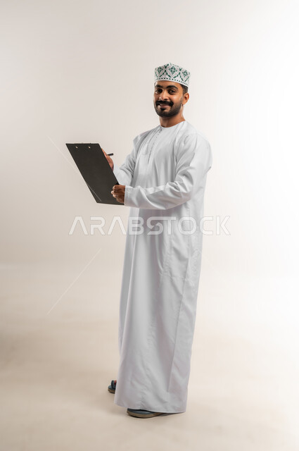 تدوين وكتابة التقارير، مراجعة نتائج الاجتماع، بورتريه لرجل عربي خليجي عماني يرتدي الدشداشة والكمة يحمل بيده كراسه تدوين الملاحظات يقوم بتصفح الاوراق، اعداد تقارير ومهام العمل، دراسة جدوى الاعمال، الجسم بالطول الكامل، خلفيه بيضاء
